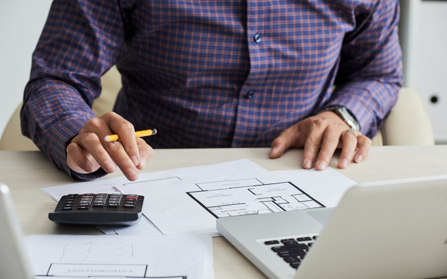 man wearing blue button down shirt, calculating 1031 exchange costs on top of building plans in front of open laptop